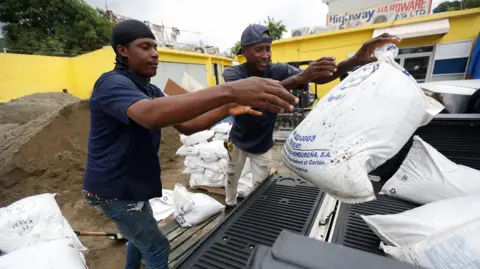 REUTERS/Octavio Jones Two men wearing t-shirts, trousers and hats throw a sandbag into the back of a pick-up truck. Piles of bags and sand are seen behind them, along with a building painted yellow and a sign that reads Highway Hardware.
