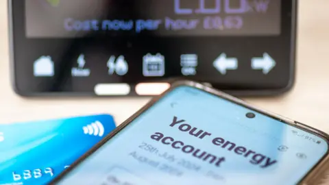 Getty Images Smartphone with a page open on somebody's energy account, sits next to a debit card and in front of a smart meter display unit. 