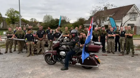 David Bathgate About a dozen military personnel, mainly men, in combat trousers surrounding the gun. There is also a man in the foreground on a vintage burgundy motorcycle