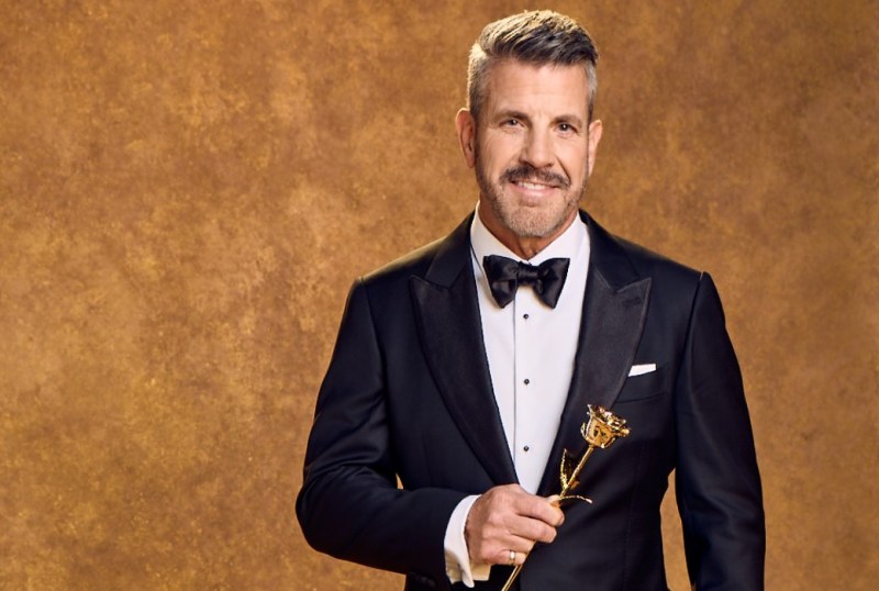 Man in a tuxedo holding a golden rose trophy, smiling against a warm, textured background. Suitable for articles about awards shows, fashion, or celebrity events in Australia.