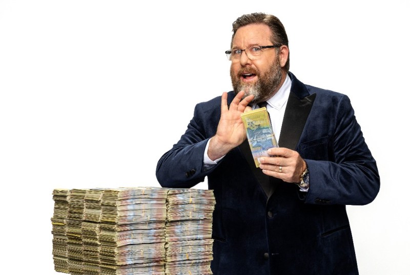 Man in a tuxedo holding Australian banknotes, standing beside a large stack of cash, promoting financial success or wealth management.