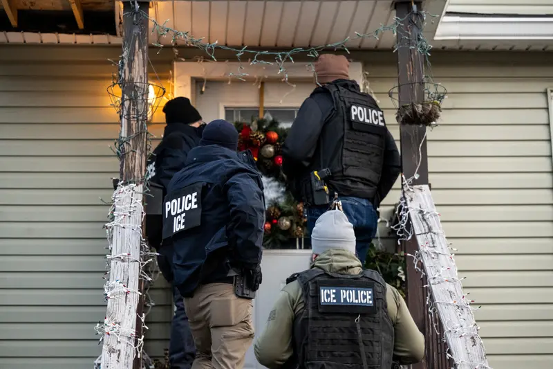 Four officers wearing tactical vests that label them as police and ICE stand on steps in front of a door with a Christmas wreath on it. Christmas lights are wrapped around the railing.