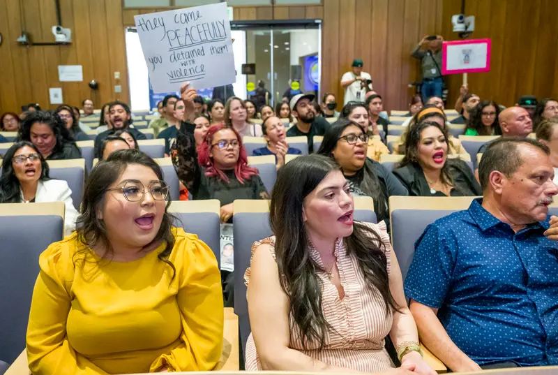 People sitting in a large room chanting. One person is holding up a sign that says “they came peacefully you detained them with violence.”