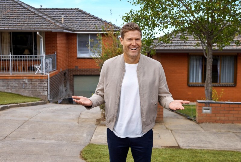 Smiling man in a casual outfit standing outside a suburban Australian home, gesturing with open hands, showcasing a welcoming atmosphere.