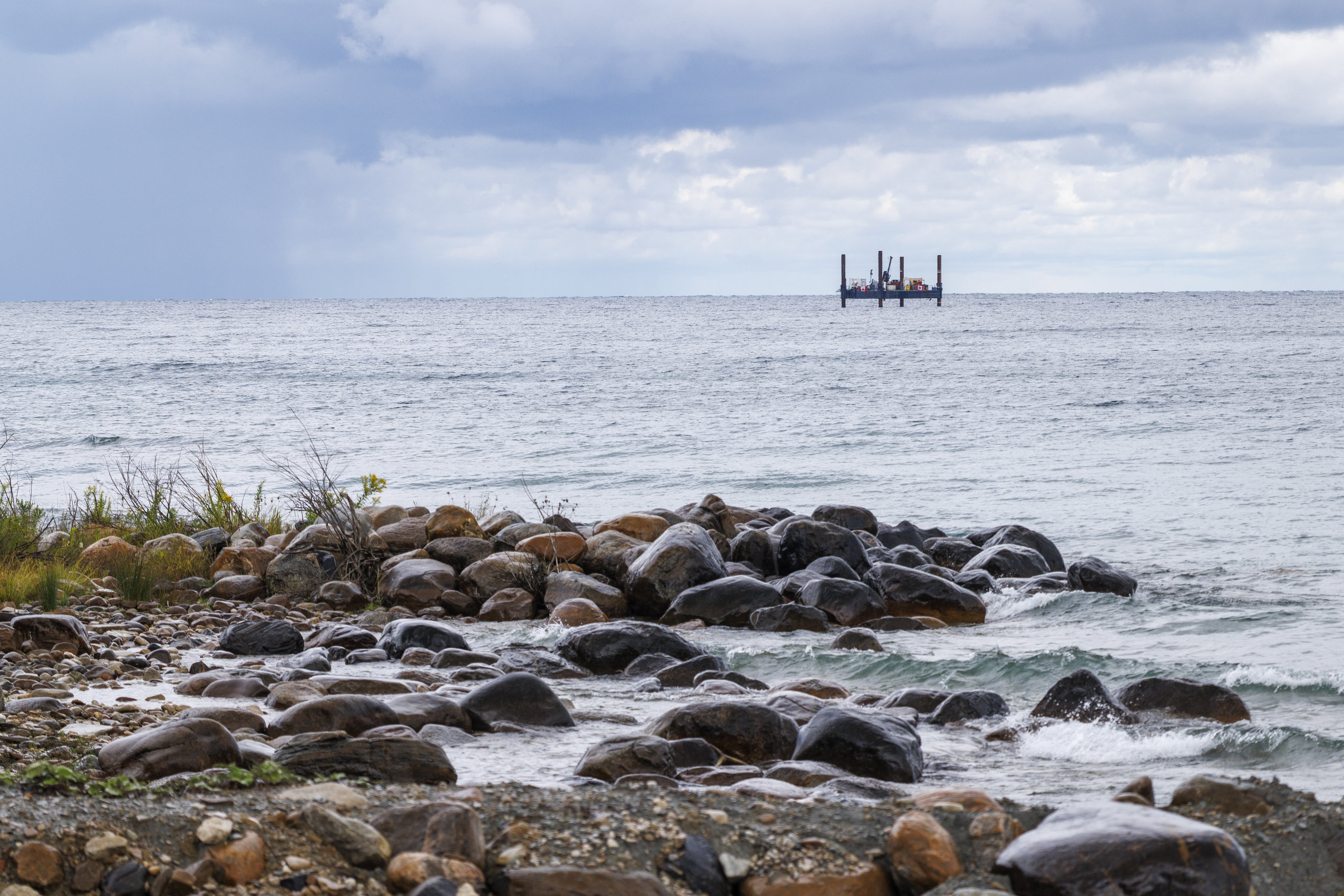 Waves lap up on the shore of Georgian Bay. In the distance, a barge-mounted drilling rig floats in Lake Huron.