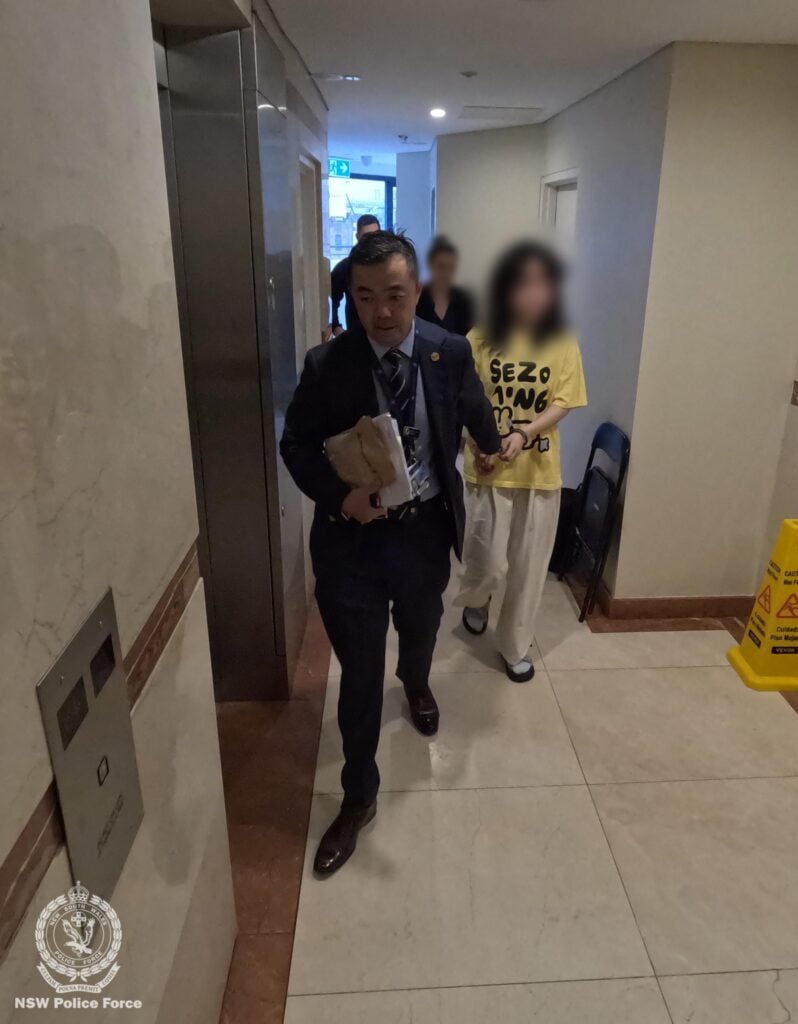 A male detective in a dark blue suit walking a woman in a yellow shirt inside a hallway