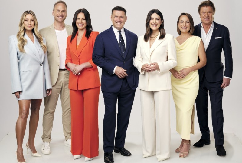 Group portrait of a diverse team of Australian presenters, showcasing a mix of professional attire in vibrant colours, including a light blue blazer, orange suit, and yellow dress, against a neutral backdrop.