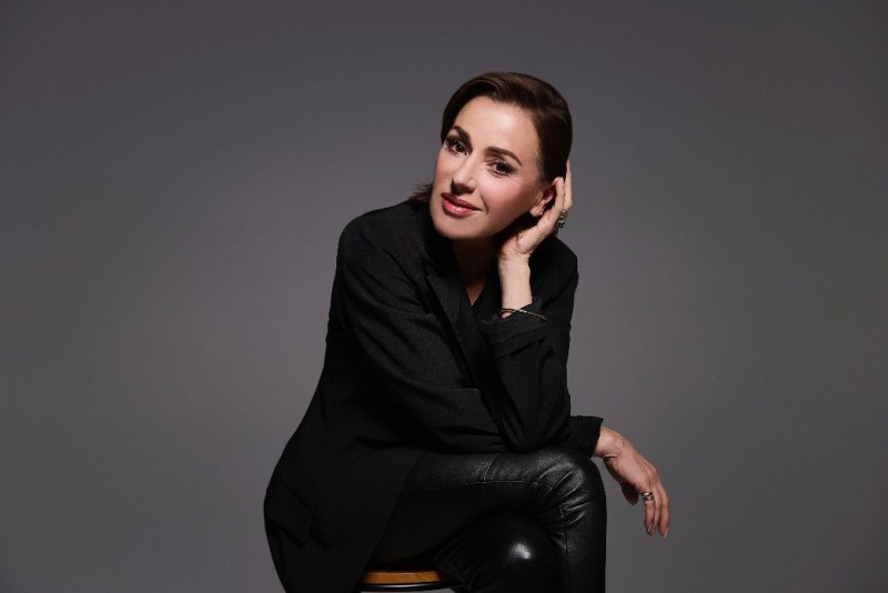 Professional portrait of a woman in a black blazer and leather pants, sitting on a stool against a grey background, showcasing a confident and stylish look.