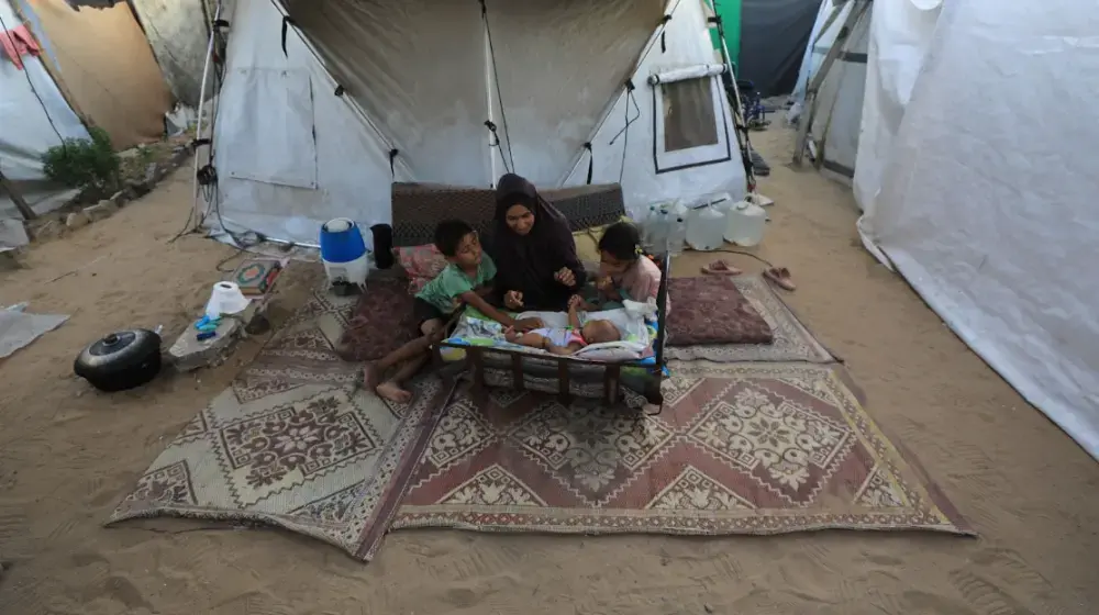 A woman sits on a small mattress beside two young children and in front of a baby in a cot, surrounded by rugs and tents in a sandy displacement camp