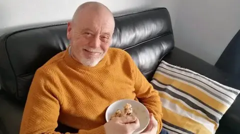 Ian Philip sits on a black leather sofa, smiling at the camera. He is wearing a mustard coloured jumper and midway through eating a bowl of cereal, which he is holding with one hand, a spoon in the other. 