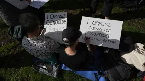 PA Media Two protesters sit on the grass writing placards 'I oppose genocide, I support Palestine Action'
