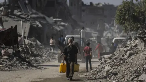 Anadolu via Getty Images A Palestinian man carries water cans among the rubble of destroyed buildings on the streets of Sheikh Radwan, Gaza City (20 October 2025)