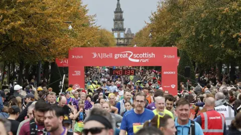 Great Scottish Run The AJ Bell Great Scottish Run, showing a large group of runners crossing the finish line under a red banner. The digital clock reads 1:40:13.9, and the scene features autumn trees and a historic building with a clock tower in the background.