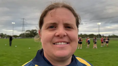 Kim Morris is standing on a rugby pitch with players behind her.  She's wearing a navy and gold training top. 