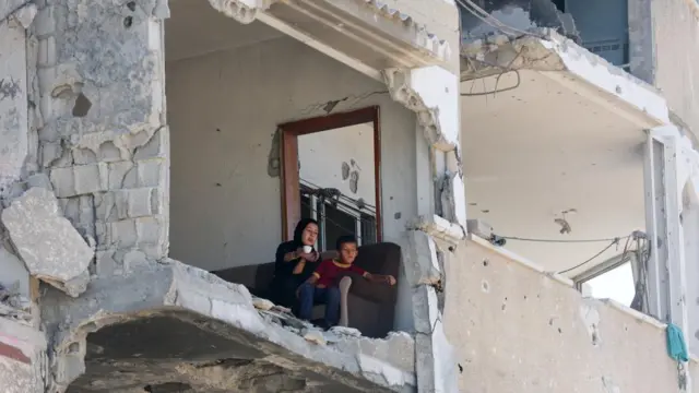 A young child and a woman sit on a sofa in a damaged building