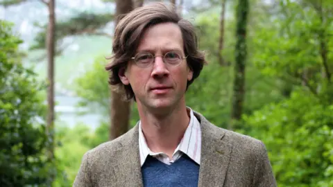 RTE A man in a brown tweed jacket, white shirt and blue jumper. He has brown hair and wears grasses. There are trees behind him.