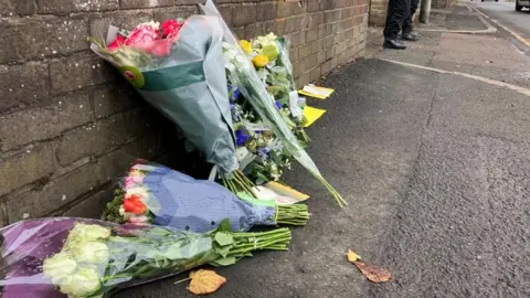 Several bunches of flowers have been leaned up against a dark brick wall on a footpath. 
