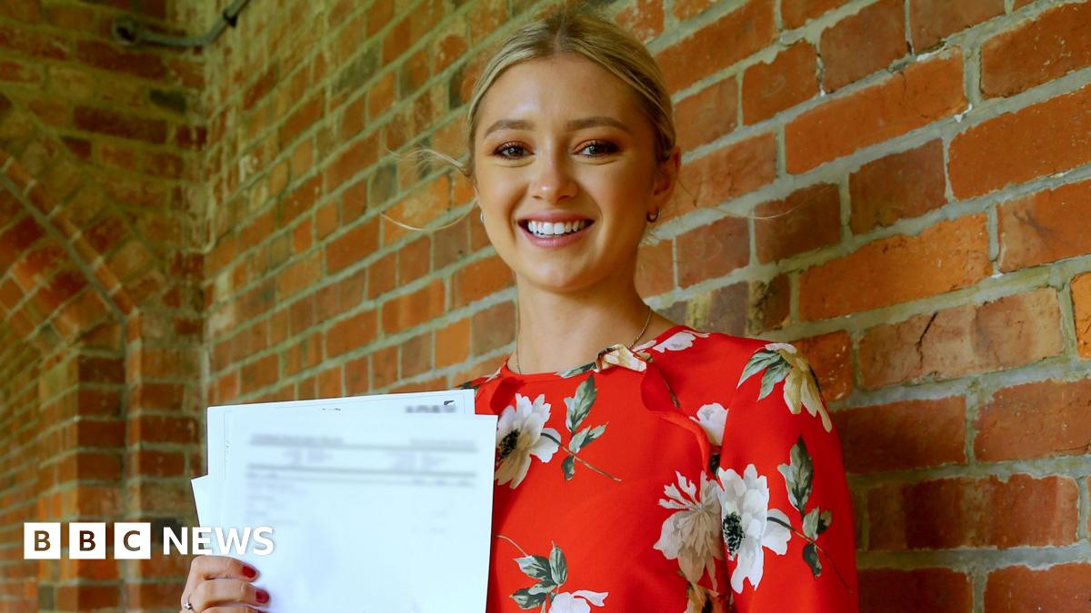 A young woman with blonde hair tied back. She is smiling at the camera and is stood in front of a brown brick wall