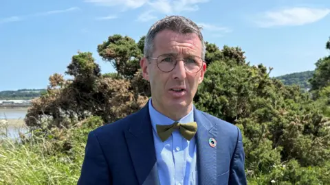PA Media Andrew Muir wears a navy suit with a blue shirt and green bow tie. He has short grey hair and wears wire-rimmed glasses. He is standing on a path surrounded by grass and shrubs. 