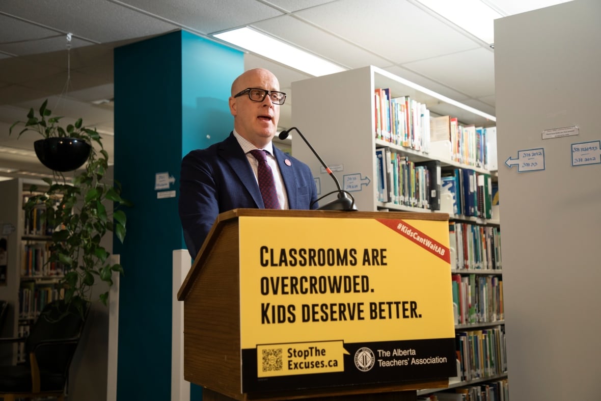 A man with a blue blazer and maroon tie stands at a lectern with a yellow sign in front, reading "classrooms are overcrowded. Kids deserve better." He is in a library.