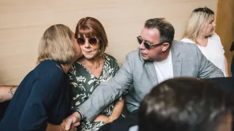 Getty Images Gisèle Pelicot sitting in Avignon Courthouse, wearing round tortoiseshell sunglasses, as her daughter Caroline leans over to kiss her on the cheek. At the same time, David, also wearing sunglasses, reaches over to hold Caroline's hand.