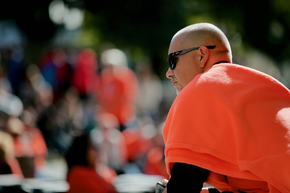 A man wearing an orange sweater is seen with a crowd behind him.