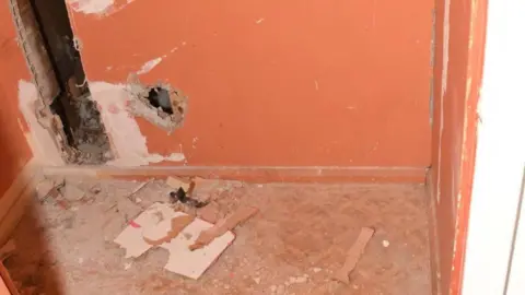 Merseyside Police An orange wall with hole and gaps and debris on the floor. The space behind the wall can be seen