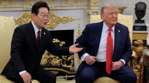 Getty Images Lee Jae Myung and Trump sitting side by side in yellow armchairs in the Oval Office. They are wearing dark coloured suits with red ties. They're looking and smiling in the same direction.