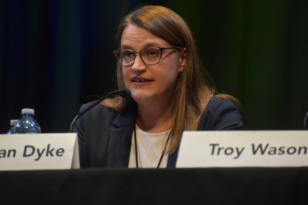 A woman with glasses and light brown hair speaks at a table with a black cloth and a dark background. 