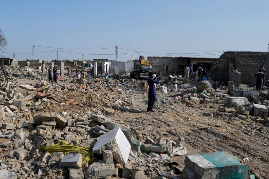 People walk through rubble as a hydraulic shovel demolishes a structure during an operation against illegal settlement of Afghan refugees conducted by local government, on the outskirts of Karachi, Pakistan on October 15, 2025.