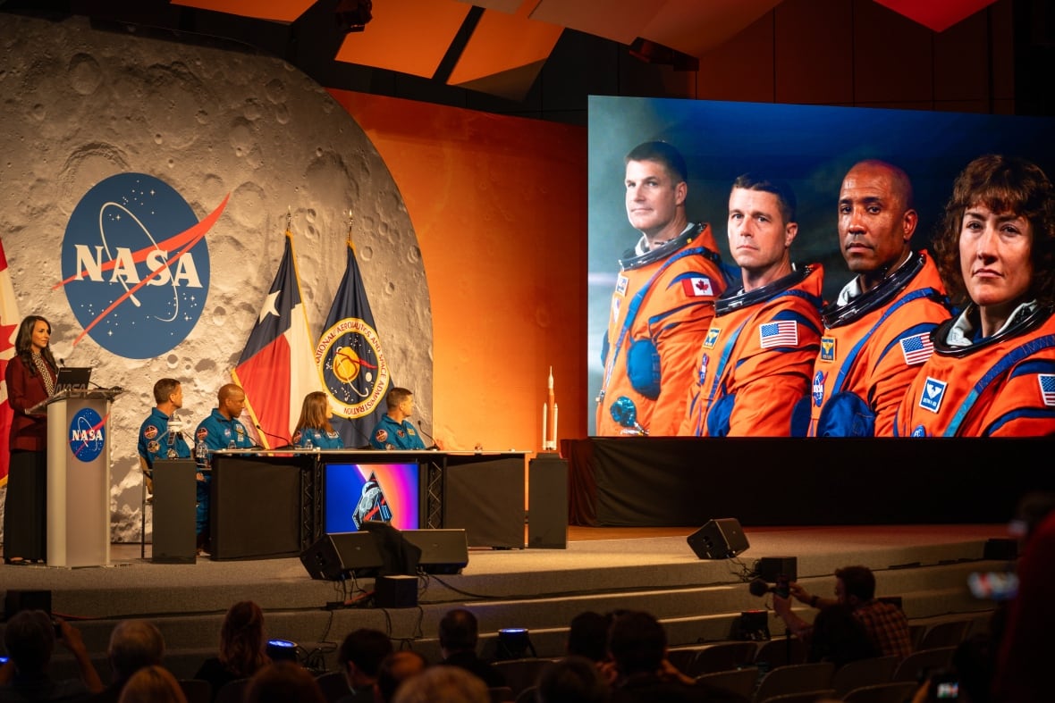 Four astronauts in blue jumpsuits sit on a stage and turn to look at a video playing of themselves in orange spacesuits.