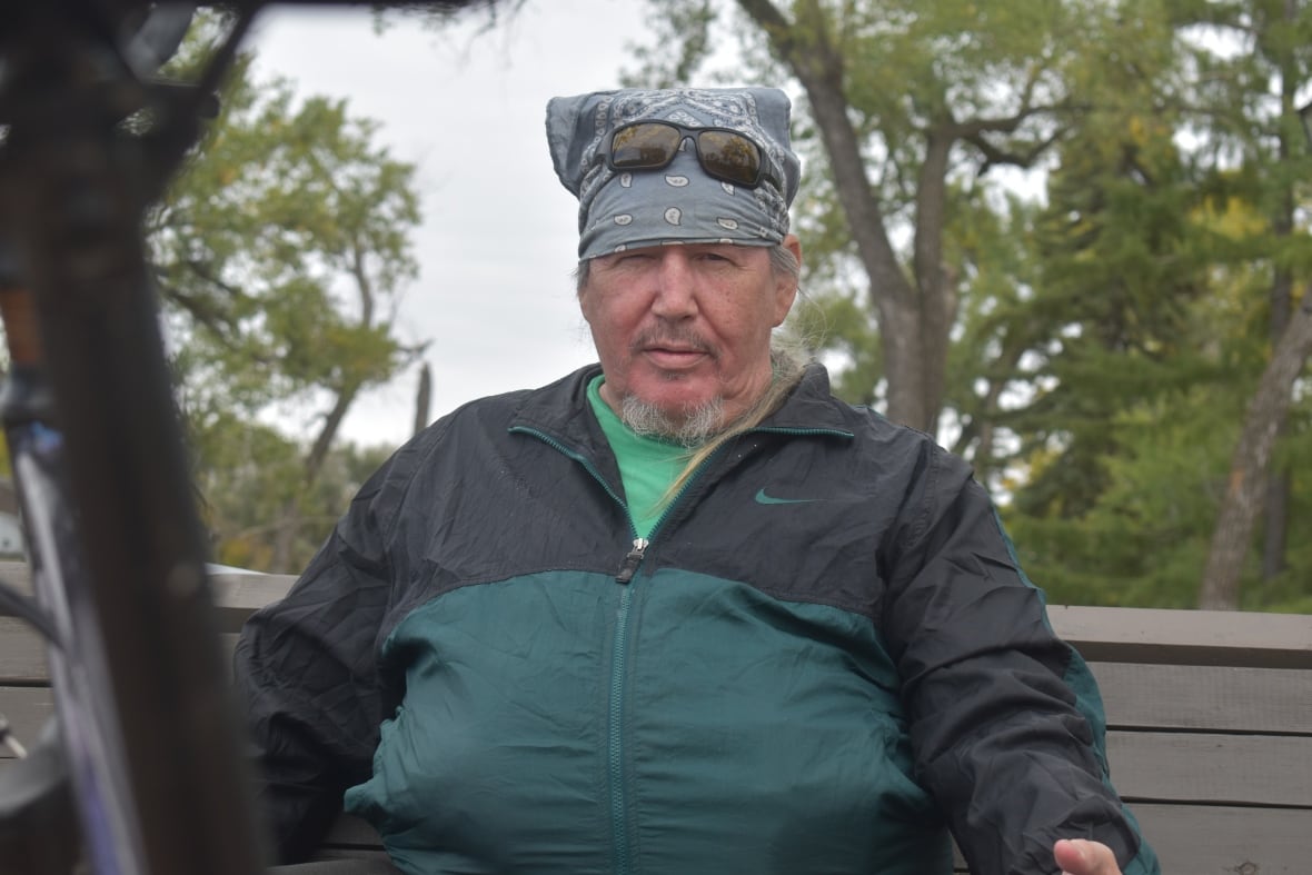 A man with a grey beard and a kerchief hat sits on a bench in a park, with a blue sky and trees in the background.