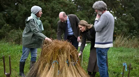 Reuters Four people gather round a stack of yellow flax in a green field. The two people in the middle in are the Prince and Princess of Wales. The prince is wearing a brown blazer, blue shirt and navy tie. The princess with long brown hair wears a brown skirt, dark green jacket and brown waistcoat. On the pair's left is a man with medium length grey hair and a short grey beard. He wears blue denim jeans and a grey sweater. On the right is a woman with short brown hair who wears a grey beanie, green waterproof coat, blue jeans and black wellington boots. 