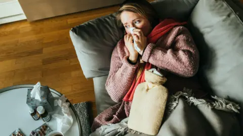 Getty Images A woman lying on a sofa while having a flu and feeling sick, with a hot water bottle on her stomach and medicine on the table