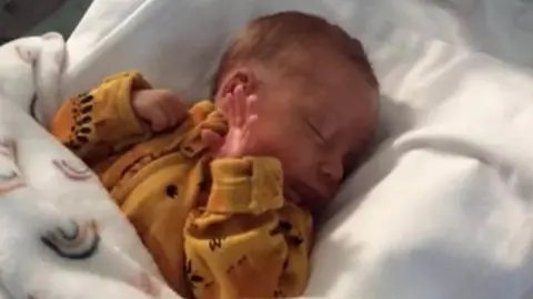 Avon and Somerset Police Brendon is pictured lying on a white sheet in an incubator. He is wearing a yellow babygrow and is covered by a blanket with rainbows on it.