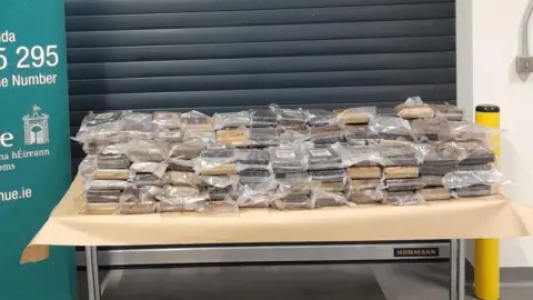 Revenue Irish Tax and Customs A close-up of a table placed in front of a grey shutter inside a non-descript room. The table has a light brown covering. On top are stacks of brown-looking material in plastic packaging, perhaps two rows with 12 stacks in each row. The drugs on the table are beside a partially-seen sign with the logo and contact details for the Irish Revenue service