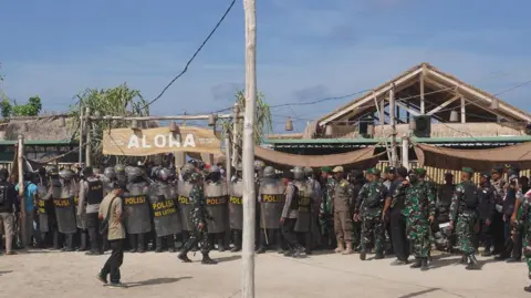 Just Finance International Police with helmets and riot shields, as well as men in green camouflge uniform,  crowd in front of a hut that has a large wooden sign that reads "Aloha".