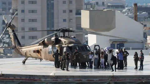 Reuters A military helicopter is on a helipad on a roof of a building. Next to it are military officials and a group of people waving and holding their arms in their air. One of them is holding a flag