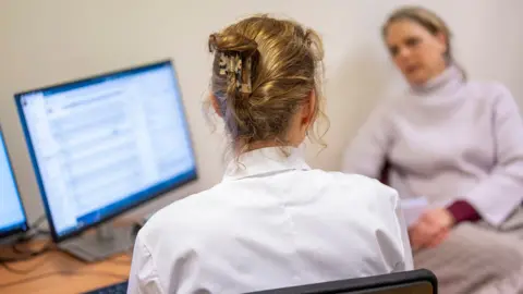Getty Images Stock photo shows the back of the head of a doctor with long blonde hair wearing a white shirt, consulting with a woman sitting opposite her in a GP office with a computer screen next to them.