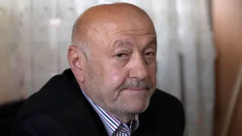 BBC Headshot of a man wearing a white shirt with red and blue stripes with a smart dark jacket. He has a greying moustache, short facial hair and is bald.