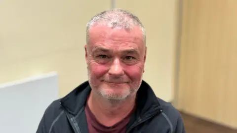 A man with thinning white hair is smiling into the camera. He has short grey stubble and is wearing a black jacket with a red t-shirt. 