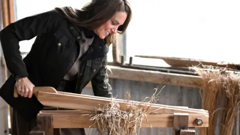 PA Media The princess operates a wooden flax machine. She has her right hand on a lever which she presses down into a flat base. In between the lever and the base is a bundle of flax. 