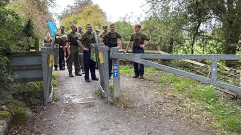 David Bathgate A group of about ten men on a countryside track standing and facing a study metal gate