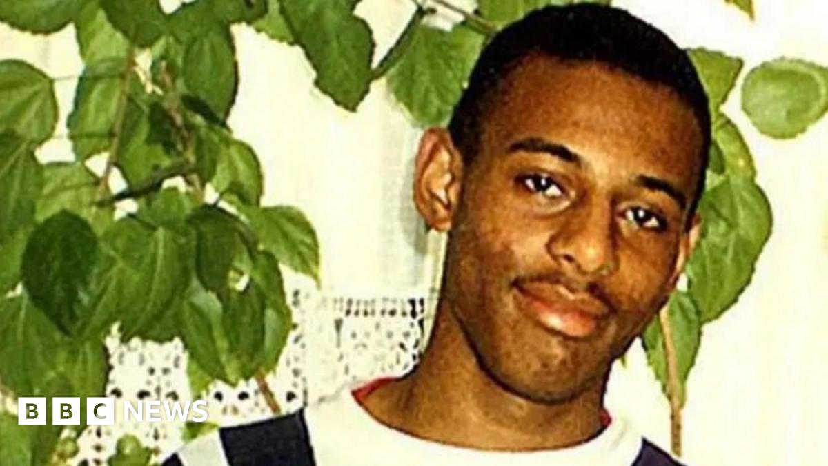 Stephen Lawrence, a young man with short black hair, wears a white t-shirt with black stripes down the shoulders. He is stood in front of a houseplant near a white wall. He is smiling.