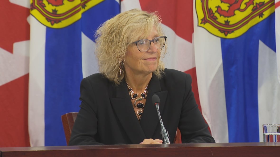 A woman in a black suit sits at a table with a microphone. Flags for Nova Scotia and Canada hang behind her.