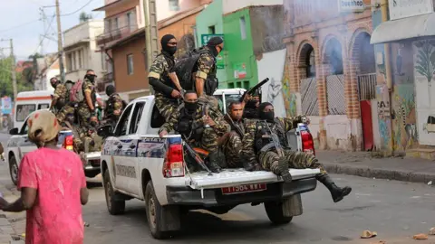 AFP via Getty Images Armed members of the security forces in military fatigues can be seen in the back of an open van driving along a road. A second vehicle with security personnel can also be seen in front of them on the road.