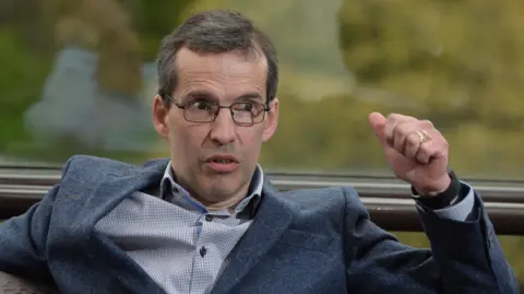 Pacemaker Press Mr Paisley wears black rectangular glasses and has short grey hair. He speaks to the right of the camera shot as he wears a dark suit jacket and a white shirt. He has brown eyes 