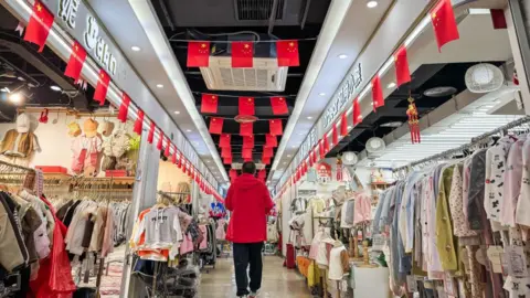 Cheng Xin/Getty Images A shopper walks through a corridor lined with children's clothing stores in a wholesale market. The shops appear quiet, while red Chinese national flags hang from the ceiling, creating a patriotic atmosphere.