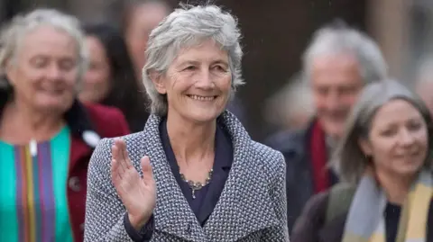 PA Media Catherine Connolly has short grey hair. She is wearing a navy suit with a blue and grey overcoat which has large lapes. She is wearing a silver necklace, is smiling and waving.