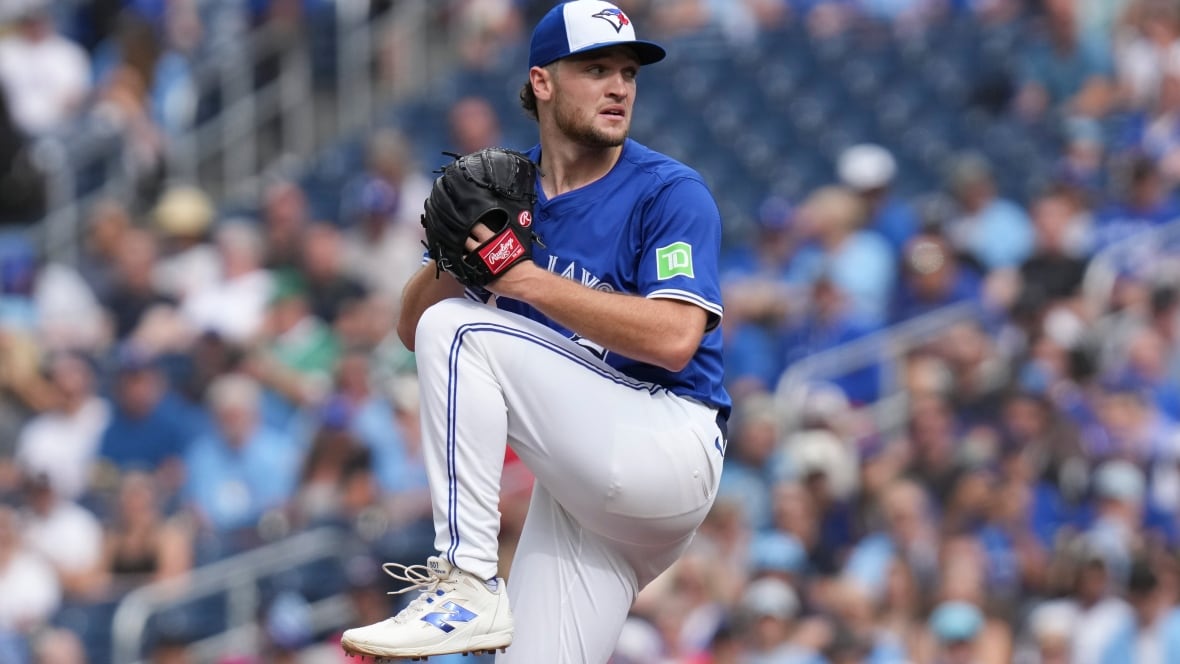 A male baseball pitcher for the Toronto Blue Jays winds up to throw the ball with his right hand as fans watch from the stands during a day game.